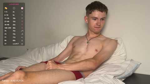 A video of a young, shirtless, fair-skinned, slim Caucasian male with short brown hair, wearing red briefs, lying on a bed with white sheets, against a plain light-colored wall.