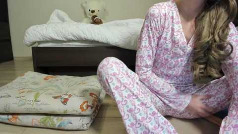 Media: Video of a woman with long, wavy blonde hair, wearing pink pajamas, sitting on a beige carpet next to a bed with a white comforter and a stuffed teddy bear.