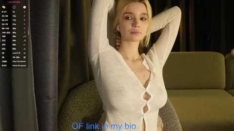 Video of a blonde woman with fair skin, medium breasts, wearing a white, long-sleeved cardigan, sitting on a green armchair. Background features dark curtains and a window. Text overlay: \"OF LINK IN MY BIG\