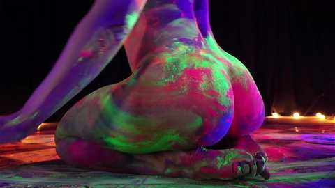Video of a nude person covered in vibrant, colorful paint, predominantly green, pink, and purple, kneeling on a floor with glowing lights in the background.