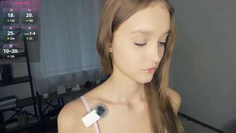 Video of a young woman with long brown hair, wearing a light pink spaghetti-strap top, standing in a dimly lit room with a digital clock displaying 18:25 and a TV screen showing a weather forecast.