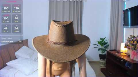 Video of a person wearing a large, straw cowboy hat, standing in a modern bedroom with a TV, flowers, and a bed.