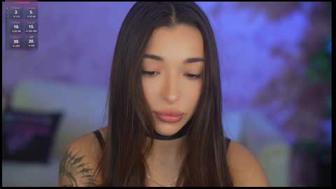 Media: Video of a young woman with long dark hair and a tattoo sleeve, wearing a black choker, looking contemplative in a dimly lit room with purple and blue ambient lighting.
