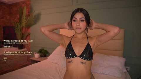 A video of a young woman with light brown skin, dark hair, and medium breasts, wearing a black glitter bikini, sitting on a bed in a dimly lit room.