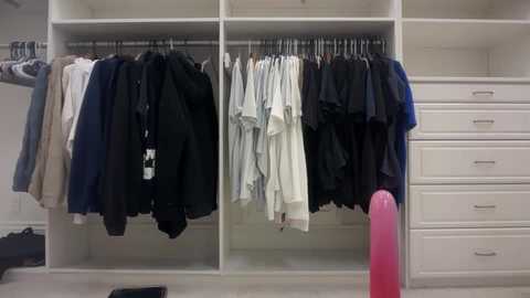 Video of a modern, minimalist closet with white shelves and drawers. Various clothes, including black, white, and navy items, hang on a rail. A pink, cylindrical object is visible in the foreground.