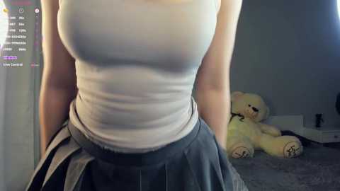 A video of a fair-skinned woman in a tight white tank top, showcasing her large breasts, and a high-waisted, pleated blue skirt. She stands in a dimly lit room with a beige wall and a plush teddy bear.