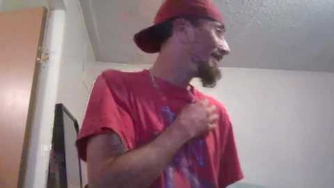 Video of a bearded man in a red hat and t-shirt, holding his shirt collar, indoors with white walls and a wooden door.