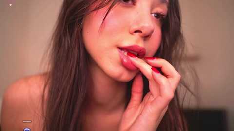 Video of a young woman with long brown hair, wearing red lipstick, seductively biting her finger in front of a blurred beige background.