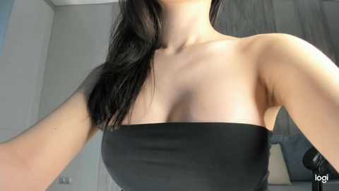 Video of a young woman with fair skin and long black hair, wearing a strapless black dress. Background includes a grey wall and a white door.