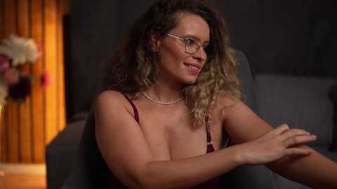 Video of a smiling, middle-aged woman with curly blonde hair, wearing glasses and a maroon bralette, seated on a dark couch. Background features dim lighting and a vase of flowers.