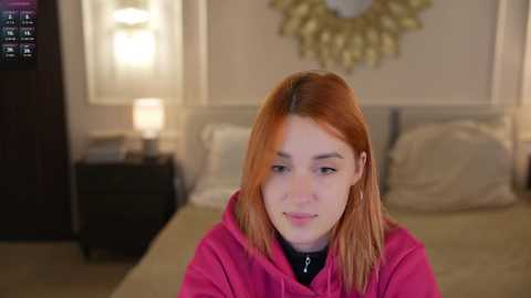 Video of a fair-skinned woman with straight, reddish-brown hair, wearing a pink hoodie, sitting on a bed with beige bedding in a dimly-lit, modern bedroom.