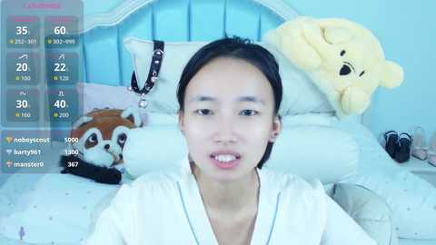 Media: Video of an Asian woman with short black hair, wearing a white robe, in a pastel-colored bedroom with plush toys.