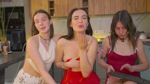 Video of three young women in a modern kitchen. They are all wearing revealing clothing, one blowing a kiss, one holding a knife, and one in a red strapless dress.