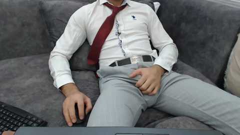 Video of a man in a white dress shirt, red tie, gray pants, and a watch, reclining on a gray sofa, typing on a laptop, with a black keyboard on the left.