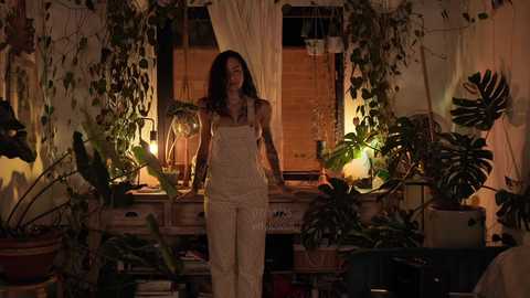 Media: Video of a woman with long, wavy hair in a cozy, dimly-lit indoor greenhouse filled with potted plants, wearing beige overalls and a sleeveless top.