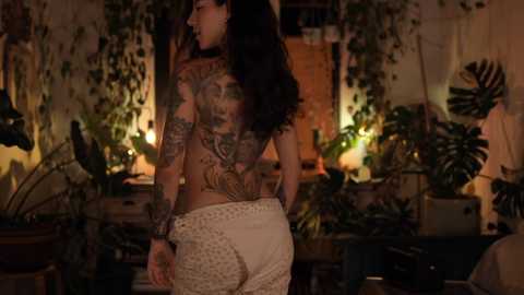Video of a tattooed woman with long dark hair, wearing a sheer top and lace skirt, standing in a dimly lit, plant-filled room with wooden furniture.