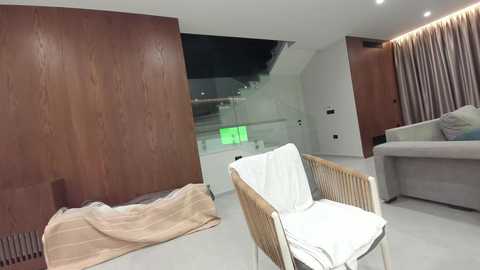 Media: Video of a modern, minimalist living room with light wood paneling, a white couch, white chairs, beige throw, and a green-lit staircase in the background.