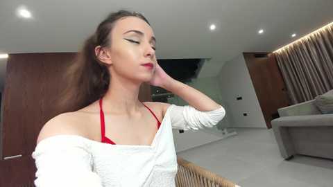 Media: Video of a slender Caucasian woman with long brown hair, wearing a white off-shoulder top and red bra, touching her neck, in a modern, minimalist living room with wooden panels and a beige sofa.