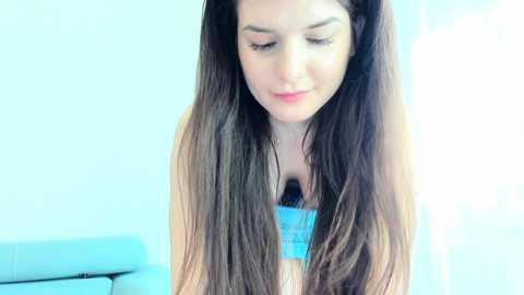 Video of a young woman with long, straight brown hair, wearing a blue top, looking down thoughtfully. Background features a blurred, light-colored wall and a hint of a blue couch.