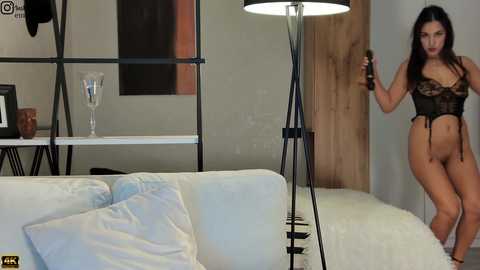 Video of a confident woman with long dark hair, wearing black lace lingerie, standing in a modern, minimalist living room with a white couch, a tall lamp, and wooden door.