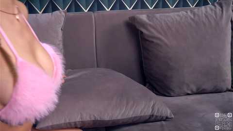 Video of a woman in pink feathered lingerie, leaning over a grey velvet couch with plush pillows, against a dark green diamond-patterned wall.