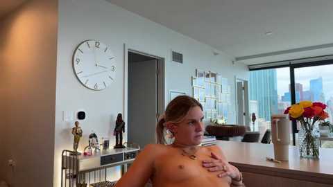 A video of a topless, fair-skinned woman with blonde hair, holding a glass of orange drink, in a modern, bright living room with a large clock and city view.