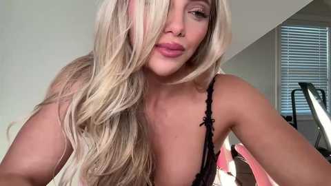 Video of a blonde woman with long hair, wearing a black lace bra, posing seductively, with a white wall and blinds in the background.