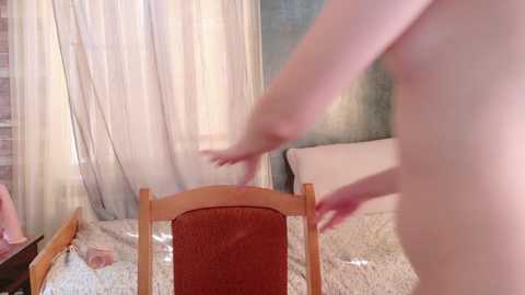 A video captures a nude woman with fair skin and a slim physique, standing in a bedroom. She is partially obscured by sheer curtains. The room features a wooden chair and a bed with white sheets.