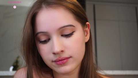 Video of a young Caucasian woman with fair skin and straight, light brown hair. She has closed eyes, with eyeliner creating a cat-eye effect. The background shows a neutral, indoor setting.