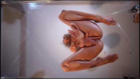 Media: Video of a nude woman with long, curly hair, standing inside a glass cube, showcasing her contortionist pose with legs spread, hands covering her breasts, and a visible vulva.