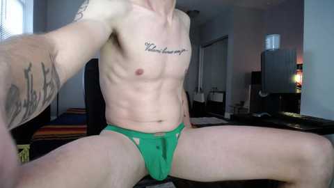 Media: Video of a shirtless, muscular man with fair skin, wearing green briefs, seated with legs spread, displaying tattoos on his arms and chest, in a modern, dimly lit room with a bed, a TV, and a water dispenser.