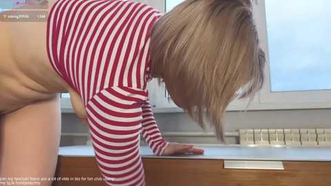 Video of a blonde woman with a red and white striped top, bending over a wooden table, showing her bare buttocks, in a modern room with a window and radiator.