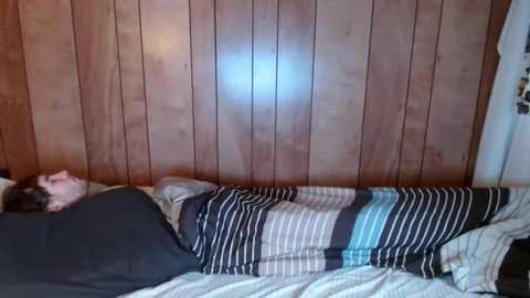 Video of a man lying in bed with a striped blanket, wooden-paneled wall, and a faint blue light in the background.