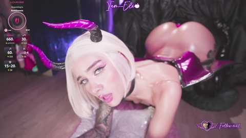 A highly stylized, digital video features a pale-skinned woman with platinum blonde hair, wearing a pink latex outfit, and black horns. She's on all fours on a black, textured surface, with a large, curved tail.
