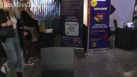 Video of a modern indoor event with a black chair, a purple banner promoting \"Moans,\" and a woman in a black jacket and jeans.