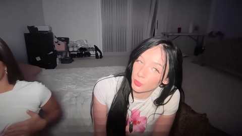 Video of a young Asian woman with long black hair, wearing a white T-shirt with pink flowers, making a funny face, in a dimly lit room with white walls and carpet.