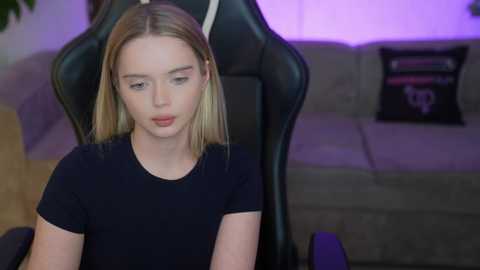 Video of a young Caucasian woman with blonde hair, fair skin, wearing a black top, seated in a black gaming chair with a grey couch and purple ambient lighting in the background.