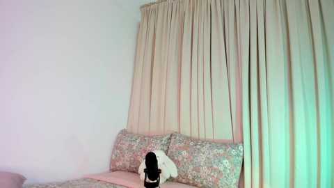 A video captures a young person with long black hair sitting on a floral-patterned sofa, partially obscured by beige curtains, against a plain white wall. The setting appears to be a dimly lit room.