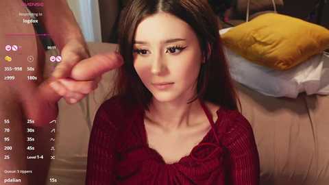 A video shows a young woman with long brown hair, wearing a red top, kneeling and touching a man's erect penis. She gazes up at him in a bedroom setting.