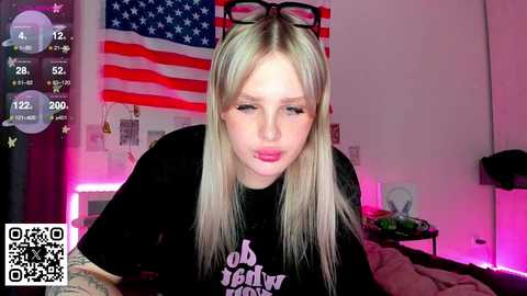 Video of a young blonde woman with glasses, wearing a black t-shirt, in a room with an American flag and pink lighting, with a QR code watermark.