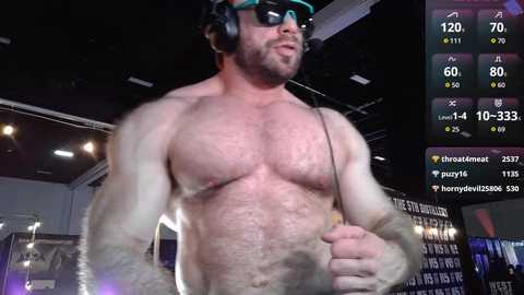 Video of a muscular, shirtless man with dark hair and sunglasses, holding a microphone, in a dimly lit room with a video game screen and stats.