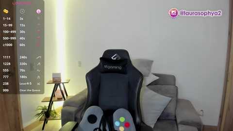 A video of a person sitting in a gray gaming chair, holding a PlayStation DualShock 4 controller, with a white wall and a wooden side table in the background.