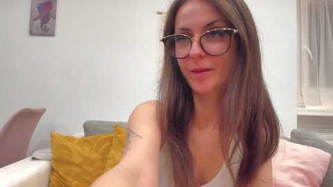 Video of a fair-skinned woman with long, straight brown hair, wearing glasses, a white tank top, and a small tattoo on her left arm. She is smiling slightly in a modern, minimalist living room with light walls and a grey sofa.