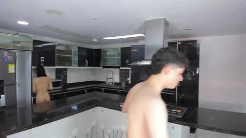 Media: Video of a modern, minimalist kitchen with a young, naked man with short, spiked hair standing at the counter, looking away. Stainless steel appliances, dark cabinets, and sleek countertops dominate the scene.
