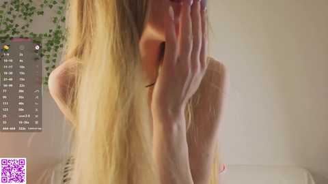 A video of a blonde woman with fair skin, hiding her face with her hands, standing in a room with a calendar and green wall decor.