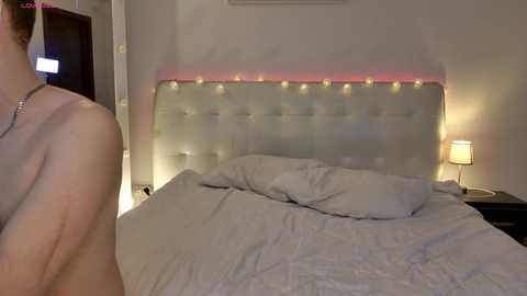 Video of a man in a bedroom, topless with a necklace, standing beside a neatly made bed with a tufted headboard, soft lighting, and a nightstand with a lamp.