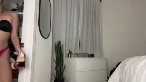 Video of a woman in black lingerie, holding a beige handbag, standing in a modern bedroom with white walls, a white dresser, and a tall plant.