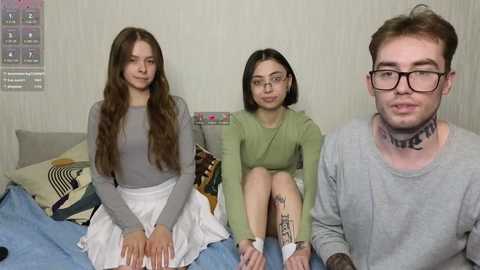 Video of three young people, two women and a man, seated on a bed with patterned bedding in a modestly decorated room. The women wear casual outfits, one with glasses, the man has a tattoo on his neck.