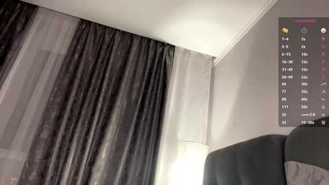 Video of a modern bedroom with a large window covered by sheer and dark gray drapes. A black upholstered bed is partially visible, and a digital clock displaying time and weather is mounted on the wall.