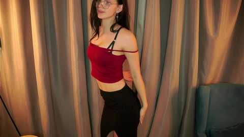 Media: Video of a slender, fair-skinned woman with long brown hair, wearing a red crop top and black pants, standing against beige curtains.